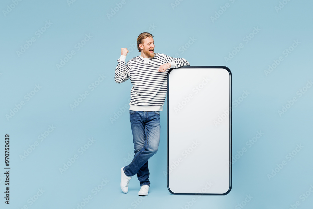 Sticker Full body young happy man he wear striped sweatshirt casual clothes big huge blank screen mobile cell phone smartphone with area do winner gesture isolated on plain blue background. Lifestyle concept.