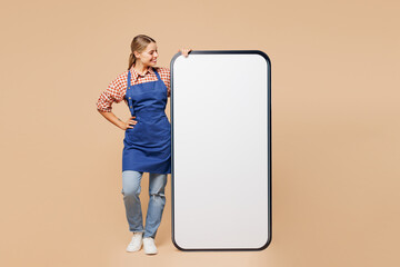 Full body young happy woman barista bartender barman wear blue apron red shirt work in coffee shop look at big huge blank screen area mobile cell phone smartphone isolated on plain beige background.