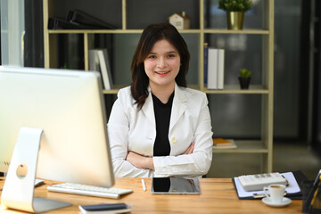Fototapeta premium Attractive young businesswoman sitting at her workplace with arms crossed and smiling