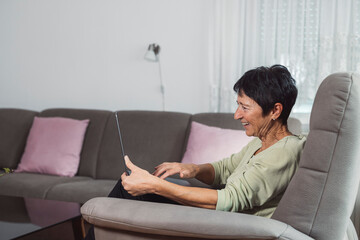 Fototapeta premium A senior woman sitting comfortably in a chair, smiling while using a laptop. The room is well-lit with a cozy atmosphere, featuring a fitness bike in the background.