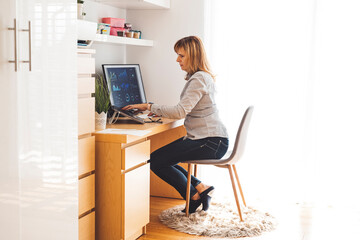 A woman working at a desk with a computer in a bright, modern home office. She is focused on her work, sitting on a chair with a soft rug underneath. The space is well-organized with shelves and plant