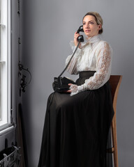 Woman in retro clothes with a old black phone before window.