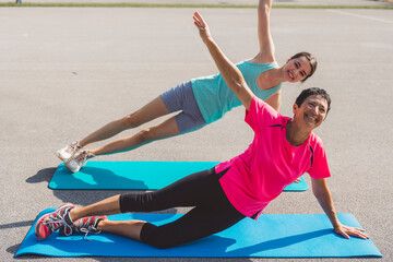 Two women exercising on yoga mats outdoors, performing side plank poses. One woman is smiling and...