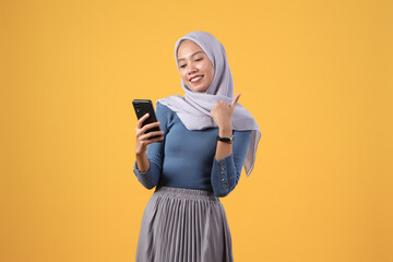 asian indonesian muslim woman holding smart phone giving thumb up finger gesture on isolated yellow background