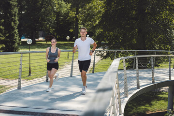 A man and a woman jogging together on a modern bridge in a park. The scene is bright and sunny, surrounded by lush green trees.