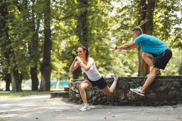 A woman performs a lunge exercise in a park while a man encourages her from behind. The scene is set in a lush green environment with trees, showcasing an active lifestyle.