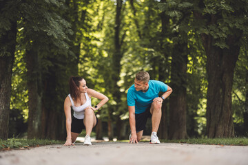 Obraz premium A man and woman in athletic attire are positioned in a starting stance on a path surrounded by trees. The woman is on her knees, while the man is in a crouched position, both ready to race. The scene