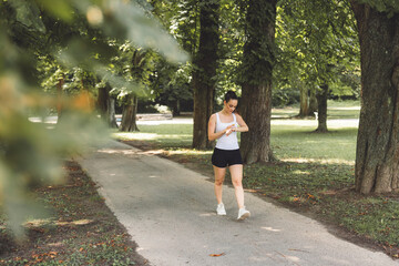 Fototapeta premium A woman in athletic wear walking on a path in a park, checking her watch. The scene is surrounded by lush green trees and grass, creating a serene atmosphere.