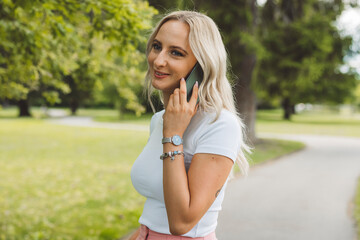 A young woman with long blonde hair is talking on her smartphone while walking in a park. She wears...