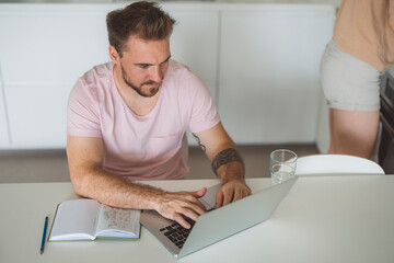 A man sitting at a table working on a laptop, with a notebook and a glass of water nearby. The setting is bright and modern, suggesting a home office or casual workspace.