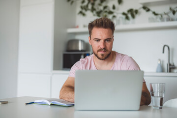 A man working on a laptop at a modern kitchen table. He has a focused expression, with a notebook and a glass of water beside him. The kitchen features plants and a minimalist design.