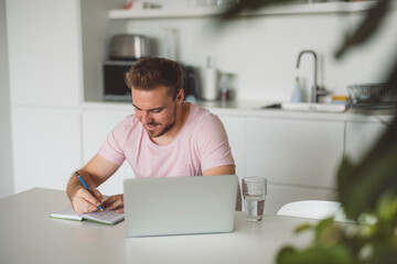 A man and a woman in a modern kitchen. The man is focused on his laptop, while the woman stands beside him holding a glass of water. The kitchen features plants and minimalist decor.