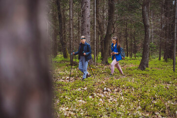Obraz premium A woman taking a photo of another woman in a forest setting. The photographer is wearing a blue vest and has long hair tied back. The subject is smiling and wearing a headlamp, standing with trekking