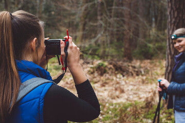 A woman taking a photo of another woman in a forest setting. The photographer is wearing a blue...