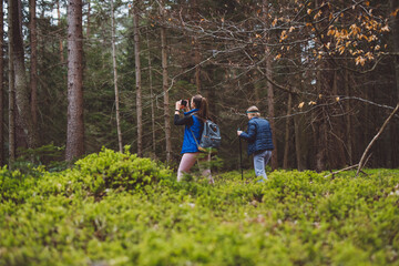Fototapeta premium Two women hiking in a forest, one using trekking poles and wearing a headlamp, the other carrying a backpack. They are surrounded by green foliage and trees.