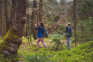 Obraz premium Two women hiking in a forest, one using trekking poles and wearing a headlamp, the other carrying a backpack. They are surrounded by green foliage and trees.