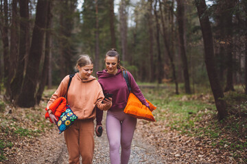 Fototapeta premium Two young women taking a selfie in a forest, smiling and enjoying their time outdoors having picnic. They are dressed in casual hoodies, surrounded by trees and greenery.