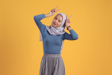 happy asian indonesian muslim woman giving victory sign finger on isolated yellow background