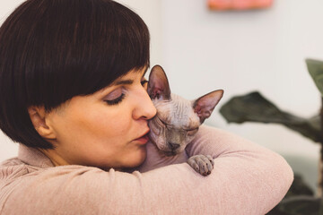 A woman with short dark hair gently kisses a Sphynx cat while holding it close. The cat appears...