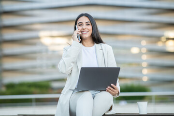 Fototapeta premium A young woman in a light suit sits on a bench, smiling while talking on her phone and using her laptop in an urban outdoor setting.