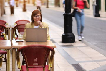 Mature stylish confident businesswoman in eyeglasses and suit works on laptop drinks coffee at European cafe outdoors. Concept of remote work from public place, digital freelance and modern lifestyle