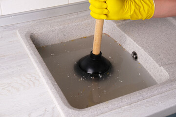 Process of unclogging kitchen sink, gloved hand holding plunger against sink background