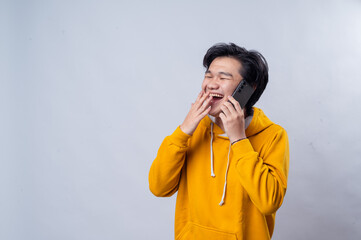 An Asian man in a yellow hoodie is laughing while talking on the phone. His hand is placed near his mouth as he smiles brightly. The background is plain white