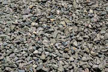 A close-up of grey gravel rocks on a sunny day. The rocks are various sizes and shapes, and they are spread out evenly across the frame