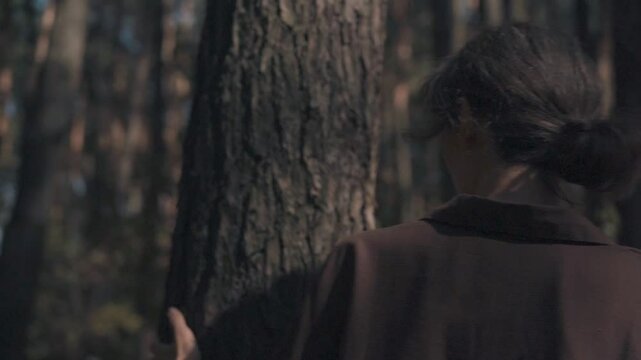 A girl of Slavic appearance in an autumn pine forest, walking around a pine tree, running her hand along the bark, leaning her forehead against the tree, looking at the crown, medium shot, slow motion