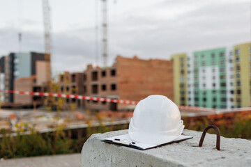 Protective white hard hat and clipboard folder of engineer on concrete build on construction site background with red-white barrier tape. Multi-storey residential and commercial real estate concept