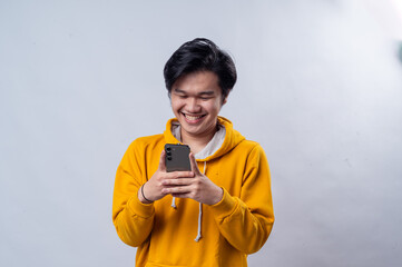 An Asian man wearing a yellow hoodie, smiling while looking down at his smartphone, texting or...