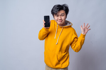 An Asian man in a yellow hoodie is holding a smartphone to his face with a surprised expression, as if reacting to something unexpected. The background is white