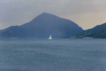 A solitary sailboat moves gently on a tranquil fjord, framed by rolling mountains as dusk approaches, creating a peaceful atmosphere.