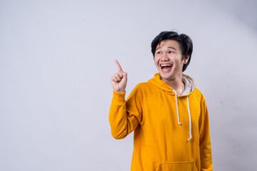 An Asian man in a yellow hoodie smiling brightly while raising his index finger as if making a point or sharing an idea. He is standing against a white background, conveying enthusiasm and excitement.
