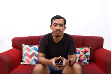 Obraz premium Funny middle age asian man sitting on the red sofa looking at the camera, doing nothing. Isolated on white