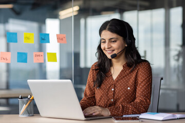 Young beautiful online support worker at workplace, hispanic woman using headset phone for video...