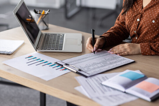 Hands of financier close-up, female accountant on paper work, fills forms, and enters data into forms, reconciles financial reports, uses calculator and laptop, writes reconciliation results.