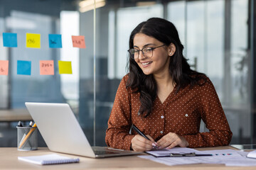 Happy woman financier doing paperwork inside office, latin american smiling pleased preparing financial report, accountant recording data and filling forms sitting at table with documents and laptop.