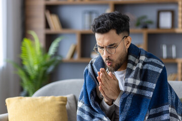 A young Indian man sits at home on the sofa covered with a blanket, rubbing and blowing air on his...