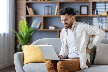 Contorted in pain, a young Indian man is at home, working on a laptop, sitting on a sofa and holding his hand behind his back