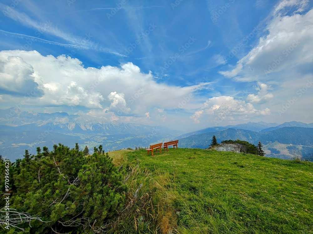 Poster Scenic view of a mountain landscape with a bench on a grassy hill under a blue sky with clouds