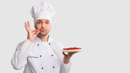Secret Culinary Recipes. Positive Chef Man Holding Baked Pie Doing Zip Your Lip Gesture Standing On White Studio Background. Panorama