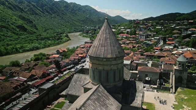 Svetitskhoveli Cathedral drone shot. Aerial view of an ancient church tower in Mtskheta, Georgia overlooking a riverside town, lush green mountains, and scenic landscapes