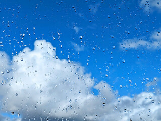 Raindrops on a window pane against a background of blue sky and fluffy white clouds