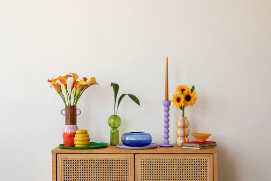Bright floral arrangement on a wooden cabinet, accented by pastel-colored vases and sculptural candleholders, adding a touch of warmth to the room. Copy space.