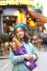 Portrait of pretty young french teenager girl in blue beret holding baguettes against the backdrop of the evening lights of a cafe or bakery on a narrow Parisian street