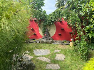 Buddha sculpture in a tranquil zen garden. A red wall with a sun motif creates a serene backdrop. Gardens Kerfouler, France