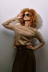 Fashion model with curly hair wearing sunglasses, posing against a light background The outfit features a neutral top and a dark skirt, showcasing modern style and elegance