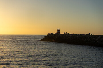 Fototapeta premium A breathtaking sunset at Nazaré, Portugal, with the sun dipping below the horizon, framed perfectly between two lighthouses at the entrance of the marina, casting golden reflections on the sea.