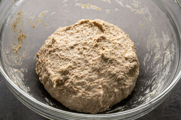 Bread dough in a glass baking bowl on a dark, neutral surface
